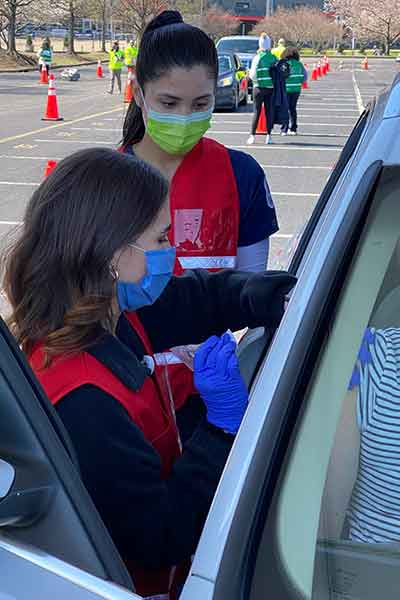 Student nurse wearing mask, vest and protective gloves gives a vaccine to a patient in a car. Behind her is another volunteer.