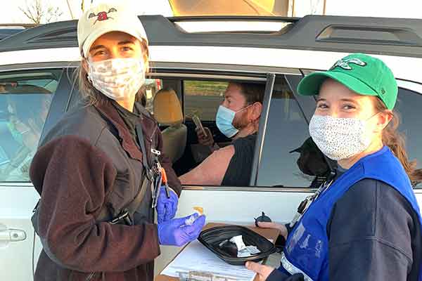 Students in masks and vests pause outside a car window before administering a vaccine