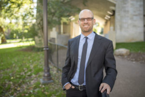 Portrait of Dr. Jonathan Metzl standing in front of a fence and building on Vanderbilt campus