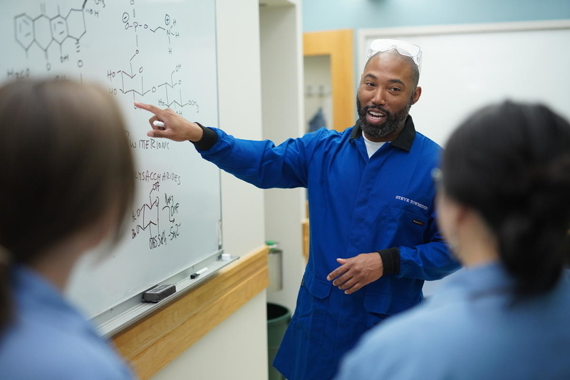 Two students watch and listen as Steve Townsend gestures to a white board containing diagrams of chemical structures