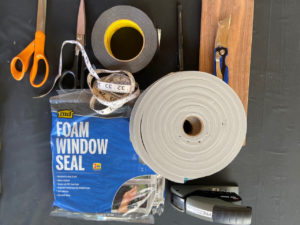 Materials and tools being used to make face masks, including scissors, measuring tape, duct tape, adhesive foam, packages of window seal, a stapler, a box cutter, and a block of wood.