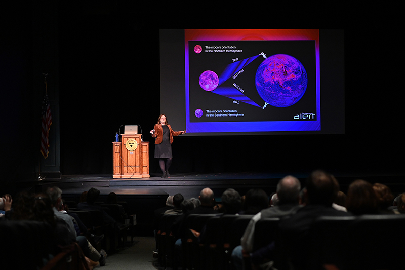 Susan Gessner Stewart stands on an auditorium stage in front of a projection screen showing diagrams of the moon.