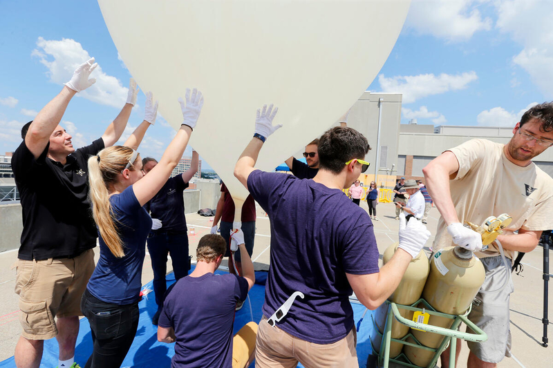 A student opens the valve on a gas tank as other students gather around an inflating weather balloon and place their hands on it to stabilize it