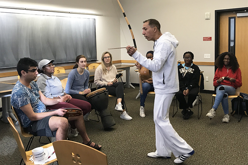 a semicircle of students sits around Gilman Whiting, dressed in capoeira uniform, as he holds capoeira tools and speaks