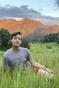 Joe Miller sitting in a grassy field with mountains and trees in background