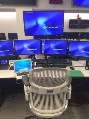 an office chair labeled "Josh Clinton" sits in front of a desk holding an array of five computer monitors, each displaying the text "NBC Universal"