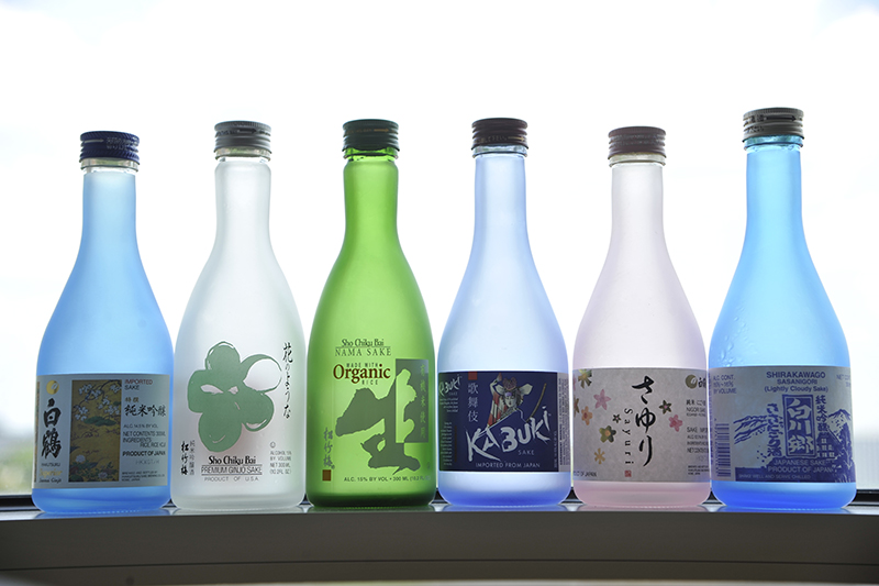A row of blue, clear, and green translucent bottles bearing labels for various brands of saki, sitting on a windowsill in the Rokas Lab