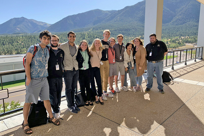 a row of students standing together on a paved terrace and smiling at the camera with tree-covered mountains in the background