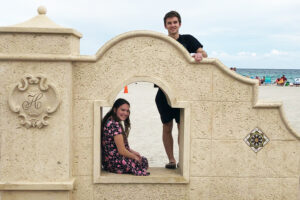 one student sits inside the archway of a stucco wall while another stands behind, with a beach and the ocean in the background