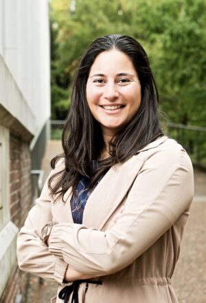 Samar Ali wearing a tan jacket, standing outside on the Vanderbilt campus with trees in the background