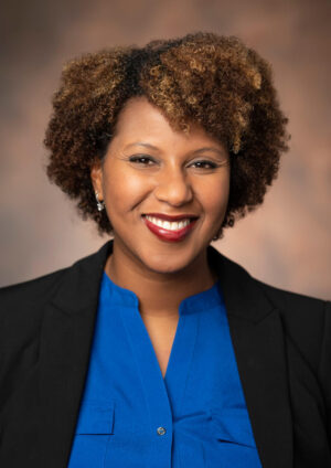 Associate Professor of Chemistry Renã Robinson, photographed wearing a blue blouse and black blazer and standing in front of a brown cloth background