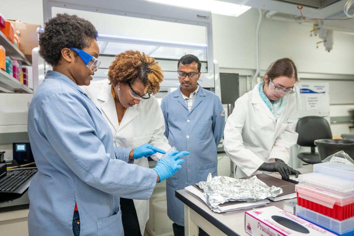 Associate Professor of Chemistry Renã Robinson, wearing a white lab coat and safety goggles, bends over to look at materials being shown to her by a student wearing a blue lab coat and safety goggles, while two other students, wearing goggles and lab coats, work in the background in Robinson's chemistry lab
