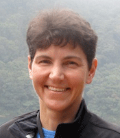 Katherine Crawford, wearing a blue shirt and black jacket, stands outside with fog and a tree-covered hillside in the background