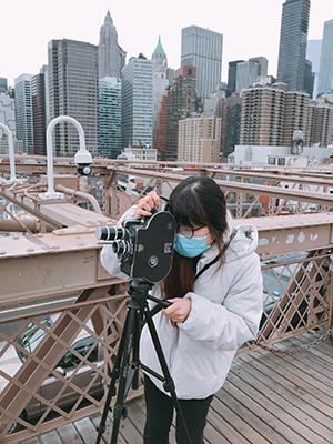 Linh Vu, wearing a tan coat and black leggings and a blue mask, looks through the viewfinder of a movie camera as she stands on a bridge with the New York City skyline in the background