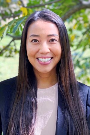 Katherine Wen wearing a navy blue blazer and white blouse, standing outside with a tree and grass in the background