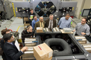 Eight men and one woman in professional attire wearing safety glasses and touring a factory space