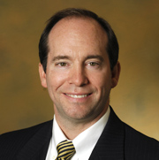 Man with short brown hair with a black and gold tie, white shirt, and black blazer smiling