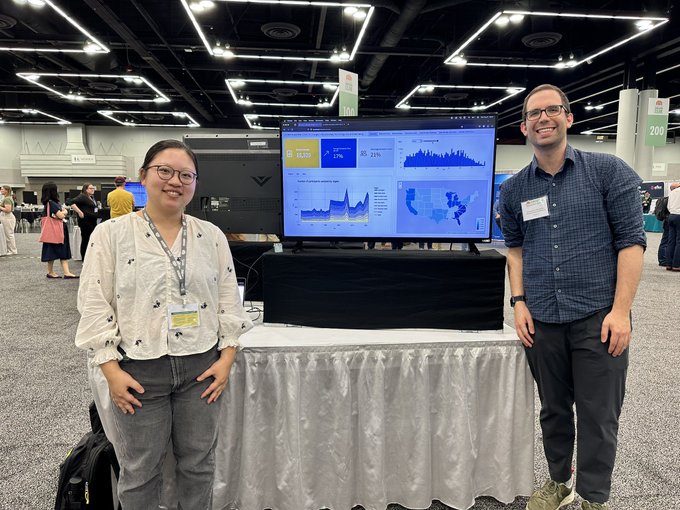 Two students (Asian woman and Caucasian man) flank digital display of their work. The screen is perched on a pedestal on top of a cloth-covered table.