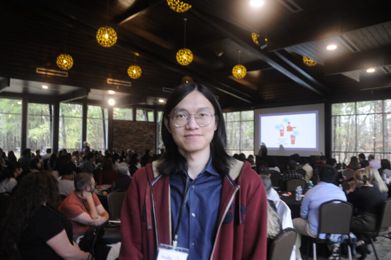 Chinese man with long hair and round wire glasses smiles at the camera; behind him, 100+ department members are looking at slides 