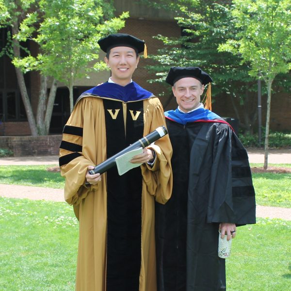 Chinese American man in gold doctoral robes and white man in black faculty robes grin at the camera